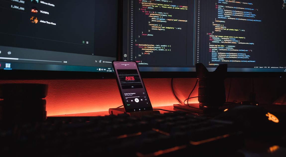 2台のディスプレイにコードが表示され、手前にはスマートフォンが置かれたデスク。株式会社クレスコが「最高のテクノロジーと絆で“わくわくする未来”を創造する」というビジョンのもと、ITサービスでビジネスニーズに応えていることを表現しています。