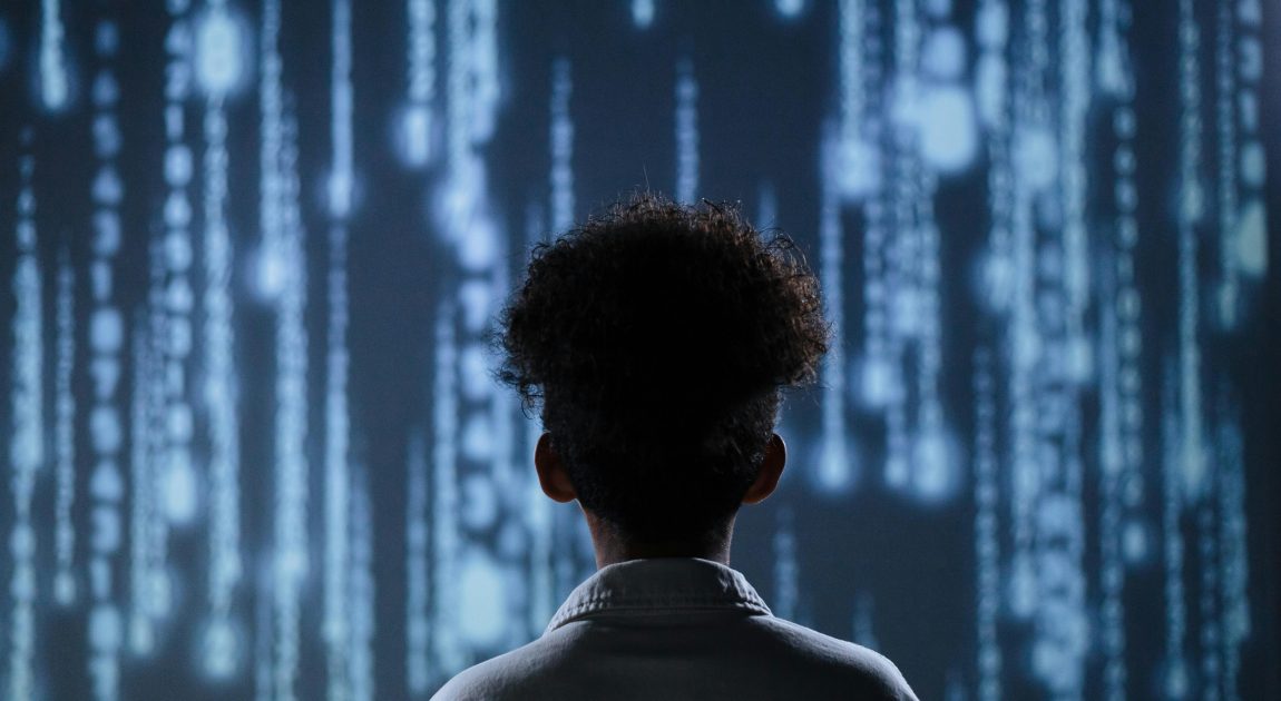 青いデジタルコードが滝のように流れる背景の前で、後姿の人物がそれをじっと見つめている。日本アイ・ビー・エムが手掛ける最先端のIT技術、特に量子コンピューター開発やAIを活用した未来のシステム構築を象徴的に表現。