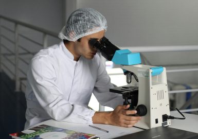 衛生キャップと白衣を着用した研究者が顕微鏡を覗き込む様子。武田薬品工業が「世界中の人々の健康と、輝かしい未来に貢献する」という存在意義のもと、革新的な医薬品の研究開発に注力し、スペシャリティ領域を強化していることを示しています。